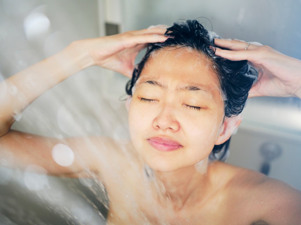 A young Japanese woman relaxing in a warm shower, eyes closed and hands in her hair.