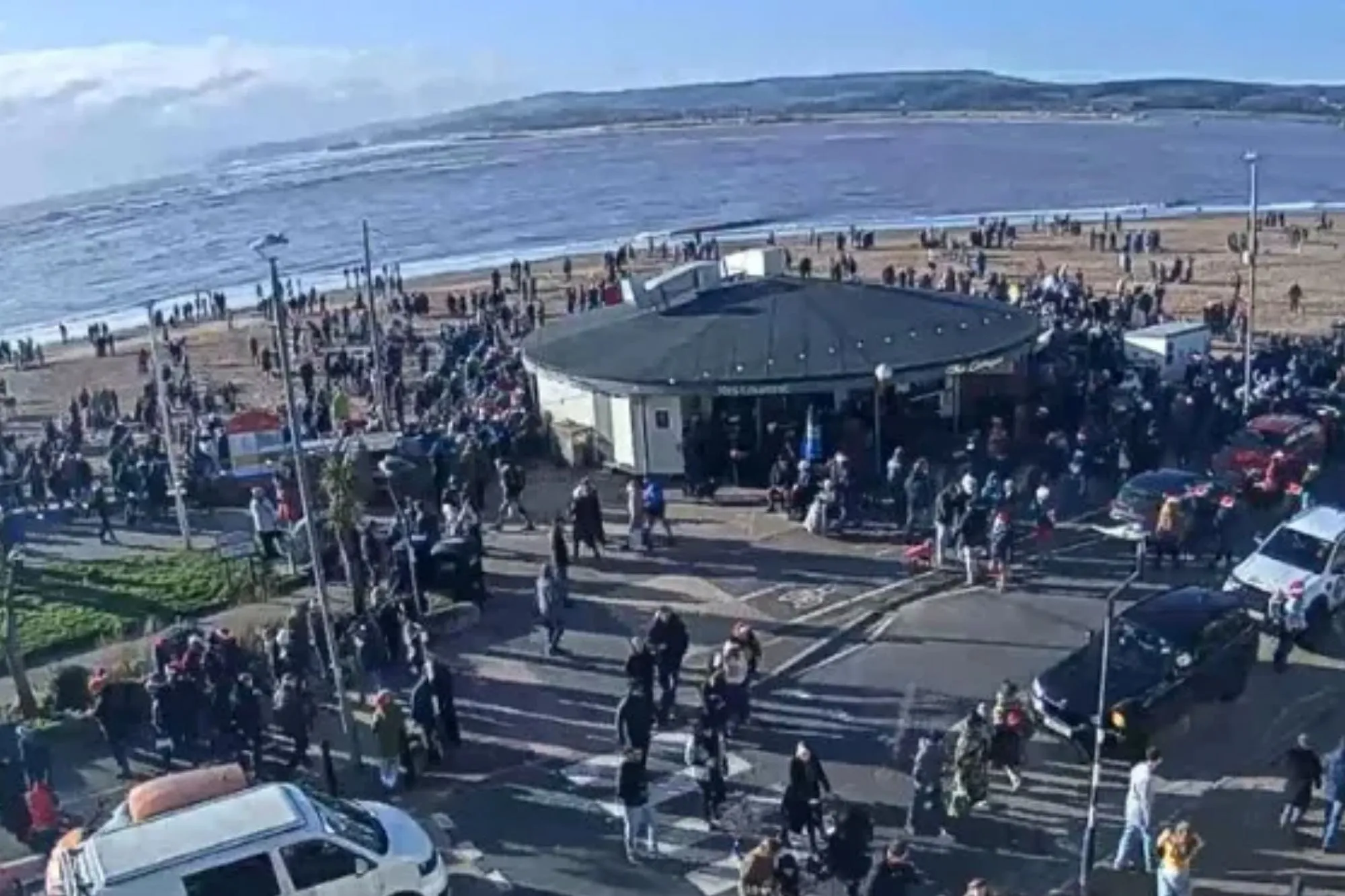 An image collage containing 1 images, Image 1 shows Large crowd gathered at Budleigh Salterton beach, where vehicles are lined up along the street