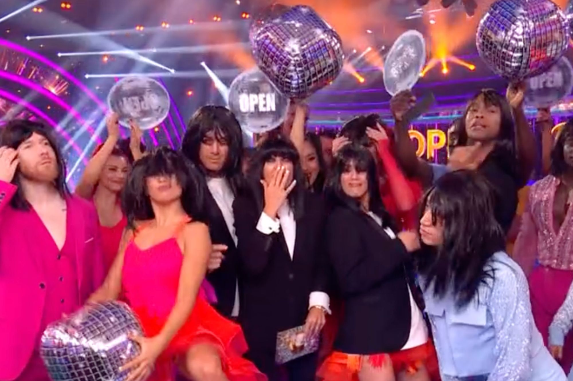 An image collage containing 1 images, Image 1 shows A group of men and women dressed in brightly colored costumes with black wigs, some holding disco balls, on a stage with a bright pink and blue background