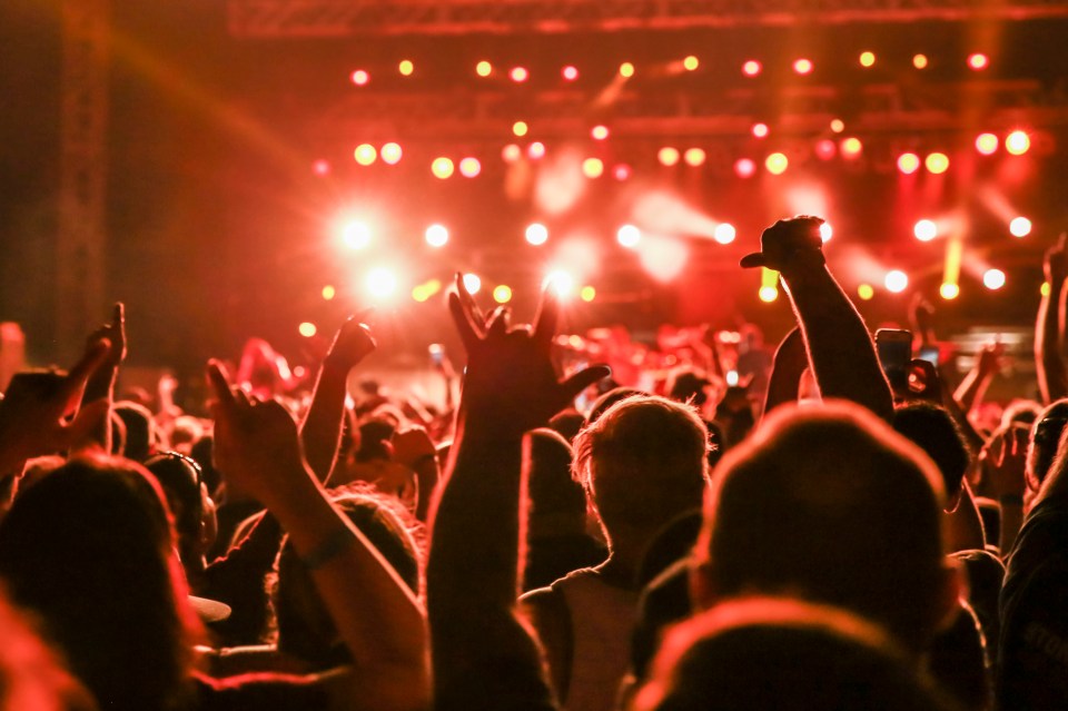 A crowd of people with their arms raised and hands in the air, silhouetted against a bright, blurry background of red and orange stage lights.