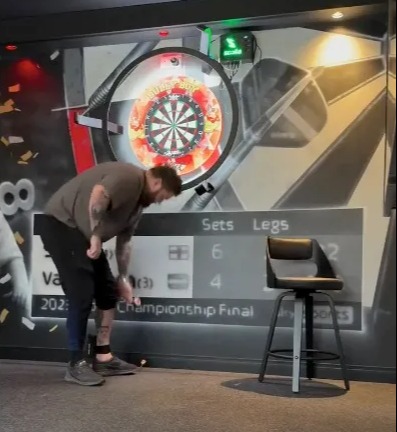 A man bending over to pick up a dart from the floor in a room with a dartboard, a bar stool, and a score display on the wall.