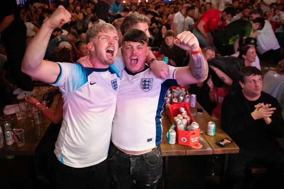 Two male England fans in white jerseys with blue details and the English crest, cheering loudly with fists raised, surrounded by a crowd at Freight Island.