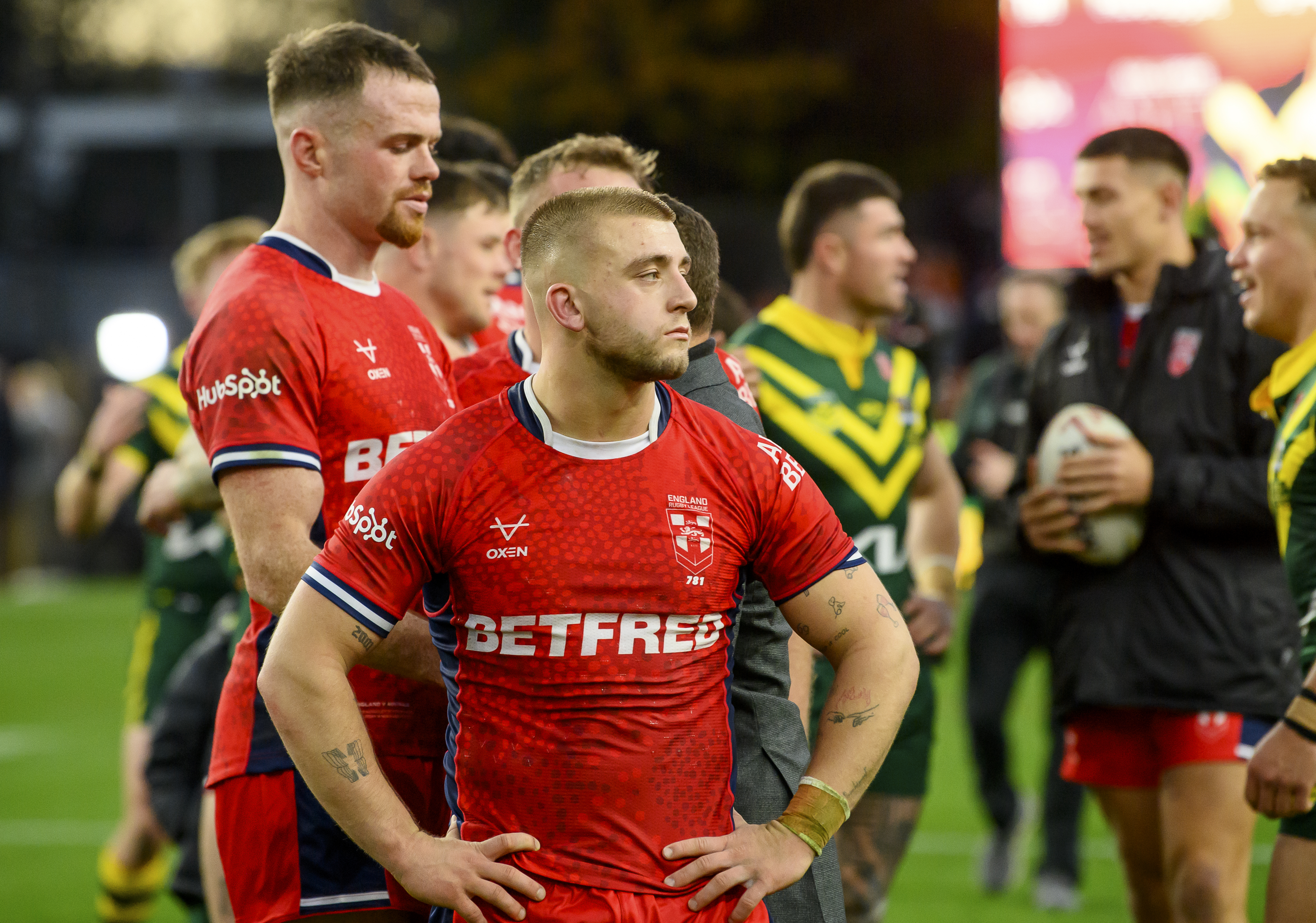 A group of Rugby League players, some wearing England red jerseys and others wearing Australia yellow and green jerseys, stand on the field.