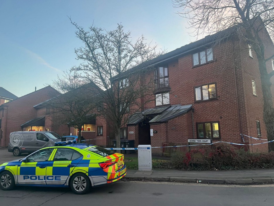 A police car parked in front of a brick building cordoned off with police tape.