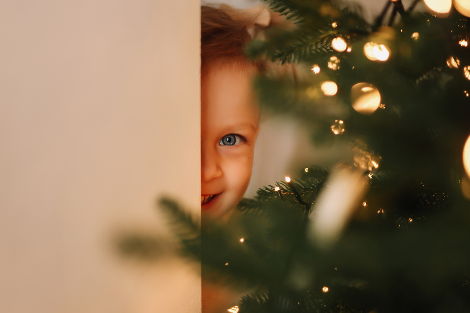 A small child with blue eyes peeking from behind a wall and a Christmas tree with lights.