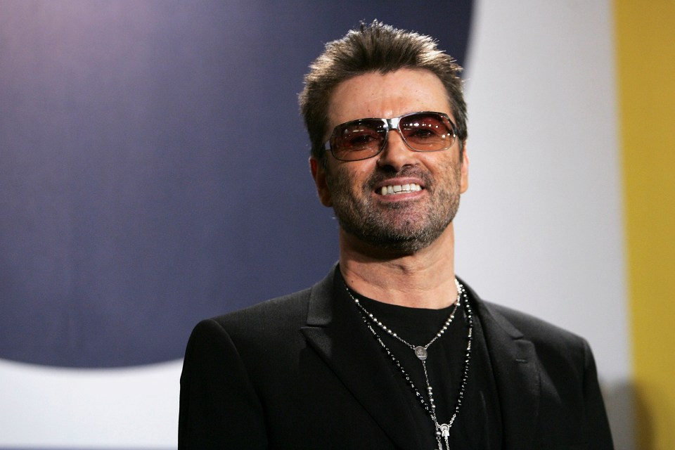 George Michael smiling and wearing sunglasses, a black shirt, and a black blazer, with a dual rosary-style necklace.
