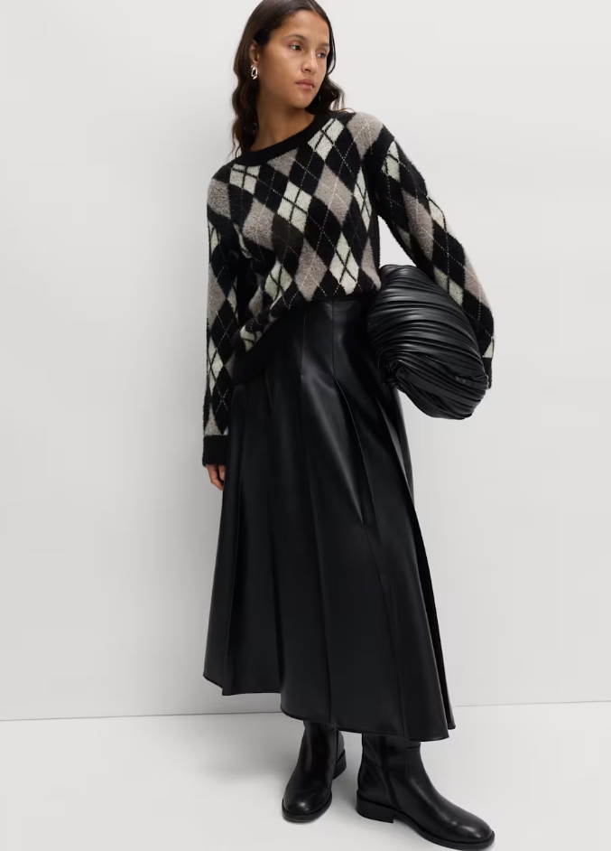 Model wearing a black and grey argyle jumper, black pleated midi skirt, black leather boots, and holding a black ruched bag.