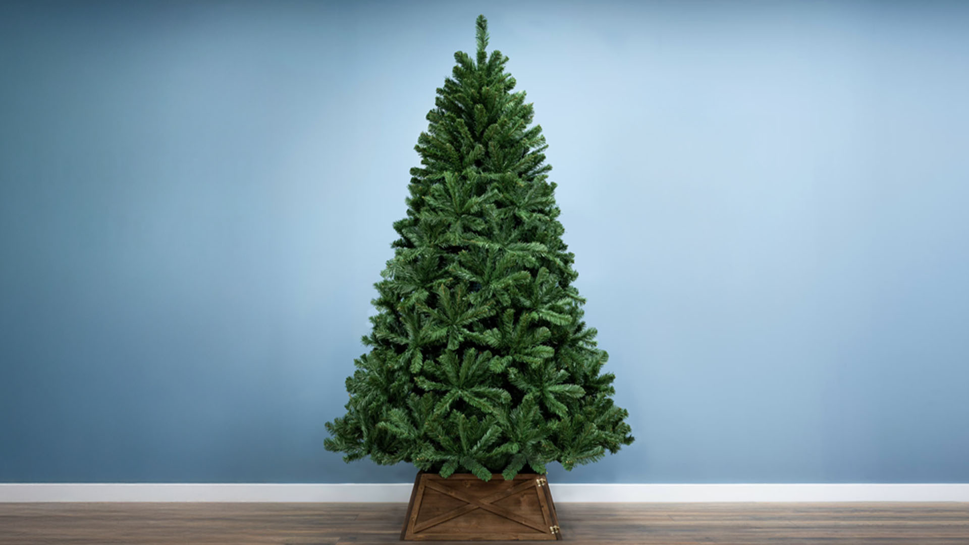 A full, unadorned artificial Christmas tree in a wooden base, against a blue wall.