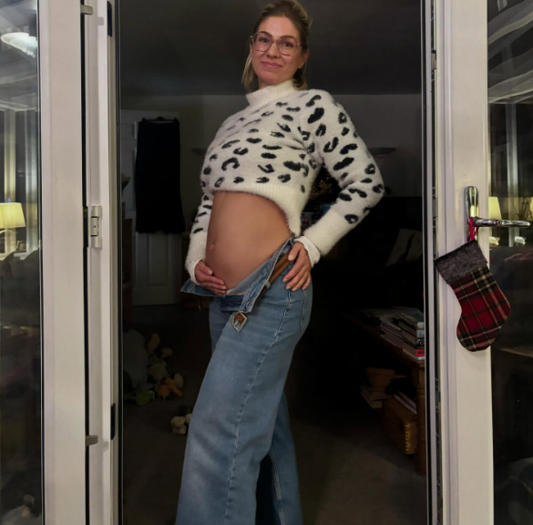 A pregnant woman in a leopard print sweater and unbuttoned jeans holds her belly, with a Christmas stocking hanging nearby.