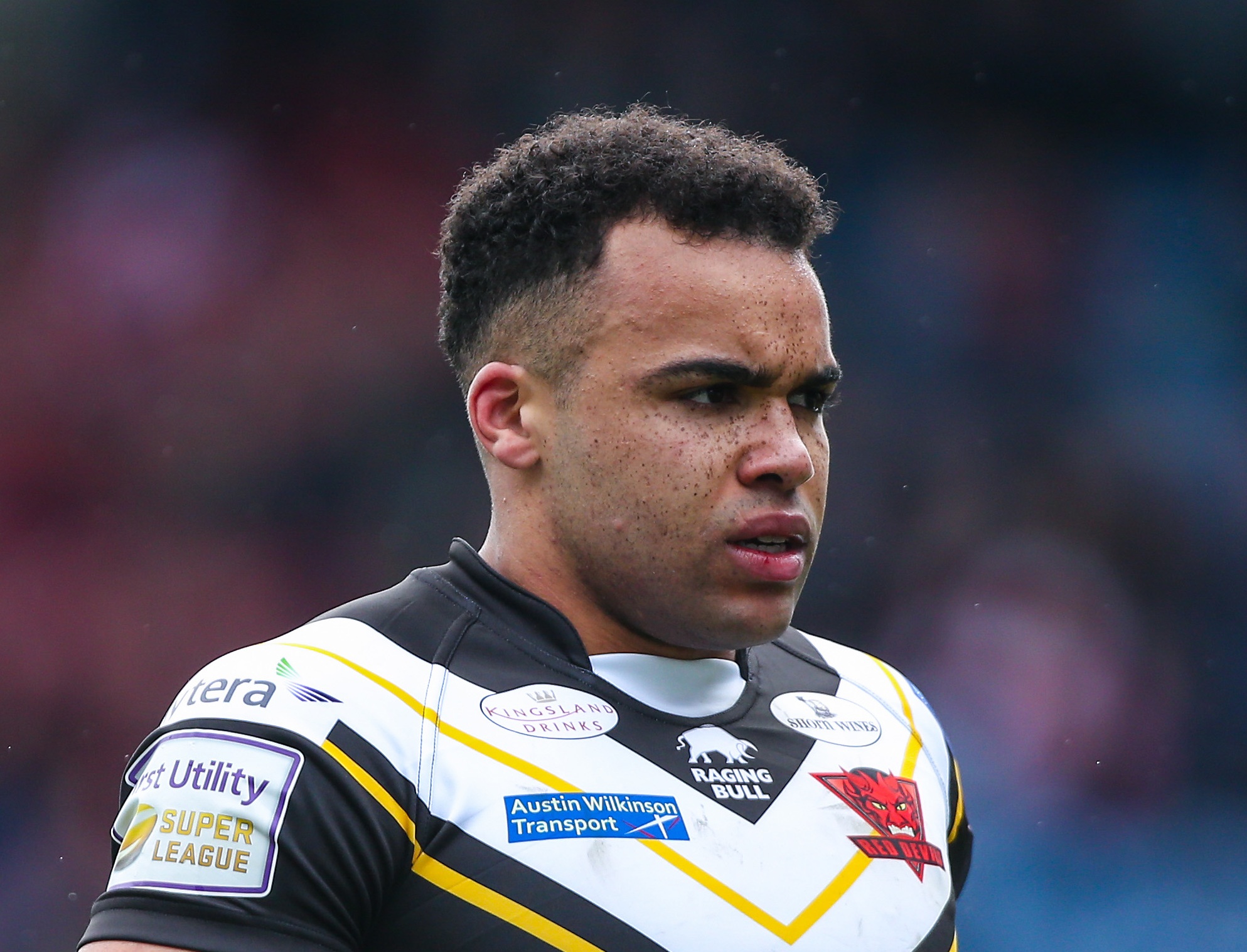 A close-up of a rugby player in a white, black, and yellow jersey with various sponsor logos, looking intently to the right.