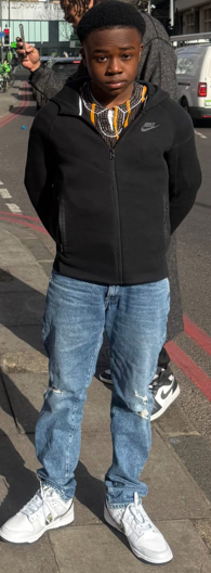 Abdul, a 13-year-old boy, wearing a black zip-up jacket, blue jeans, and white sneakers, stands on a sidewalk.