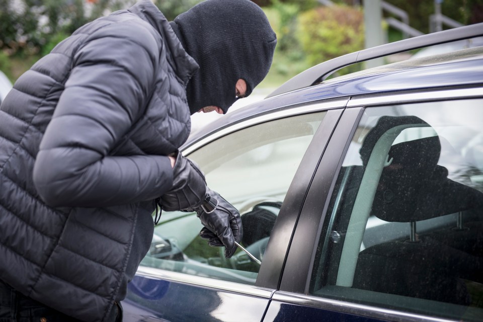 A person wearing a black balaclava and gloves using a screwdriver to break into a blue car.