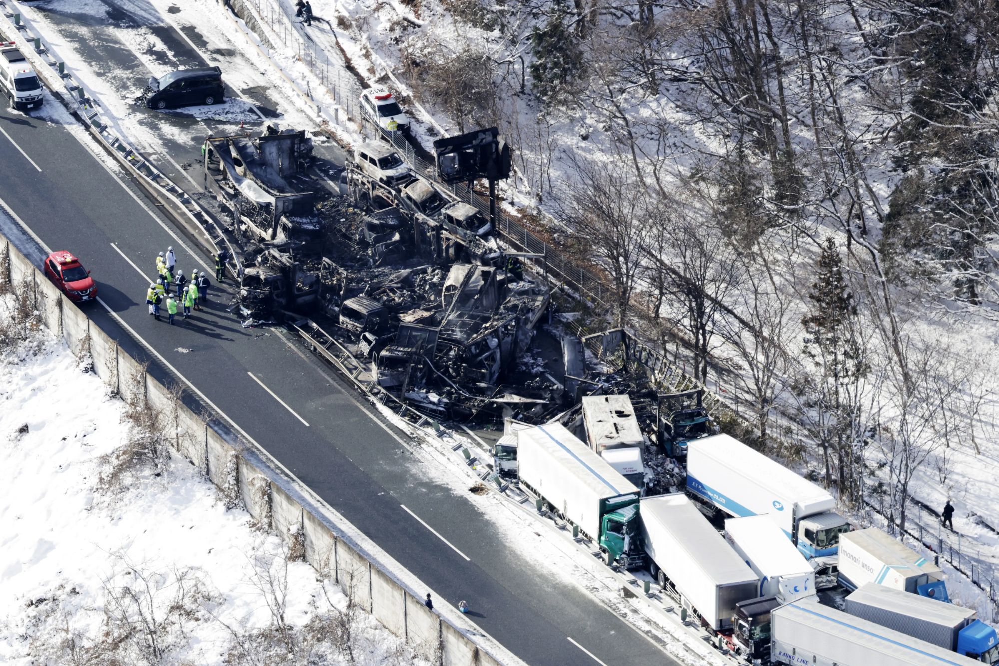 An image collage containing 1 images, Image 1 shows Aerial view of a highway pile-up involving numerous wrecked cars and trucks, some charred, on a snow-covered road with emergency responders investigating