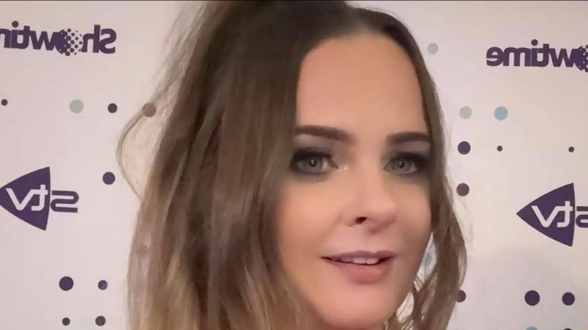 Woman with brown hair and eye makeup smiling at the camera, with a branded backdrop.