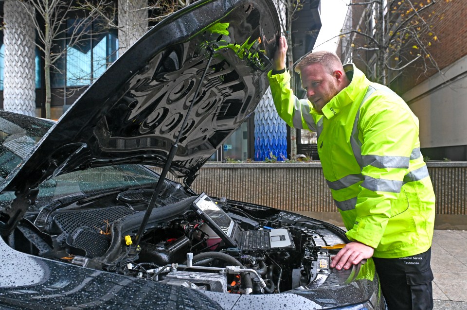 A technician in a high-visibility jacket inspecting the engine of a car with its hood open.