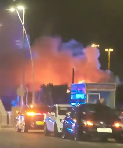 Cars parked on a street at night with a large fire burning in the background.