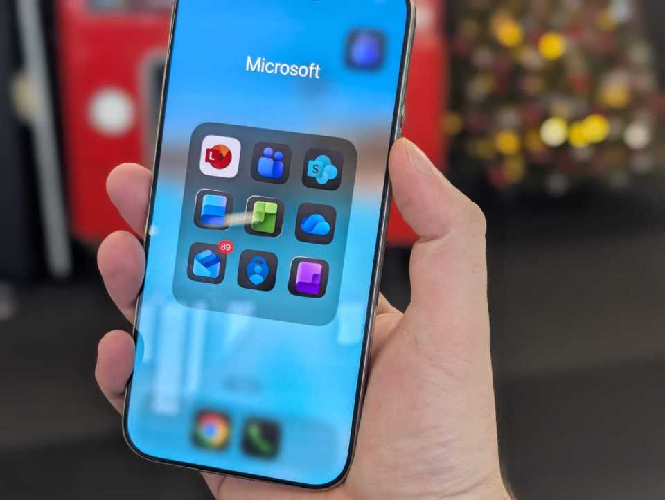 A hand holding a smartphone displaying a folder with Microsoft apps.
