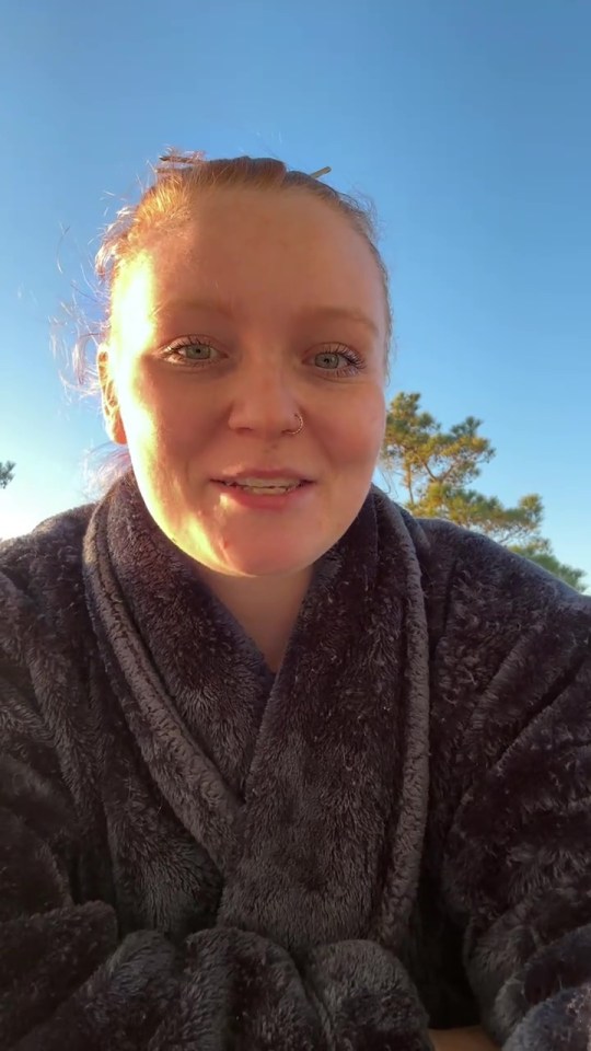 A person with red hair, green eyes, and a nose ring wearing a dark gray robe, looking up at the camera.