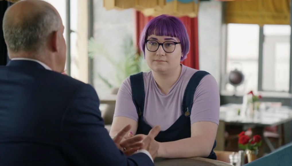 A young woman with purple hair and glasses listens to a man in a suit.