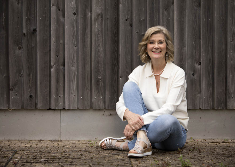 Woman in a white sweater and blue jeans sitting against a wooden wall.