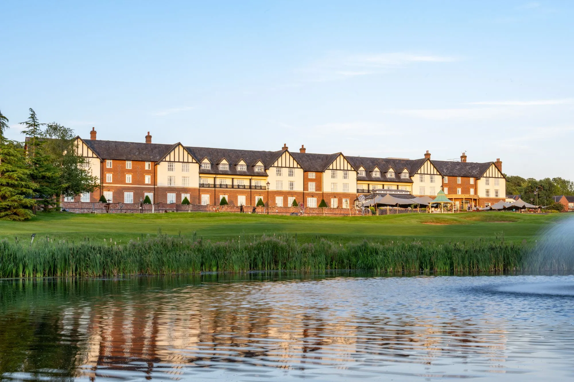 An image collage containing 1 images, Image 1 shows Carden Park Hotel reflected in a pond with a fountain