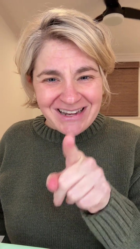 A smiling woman with blonde hair wearing a green sweater points her finger at the viewer.