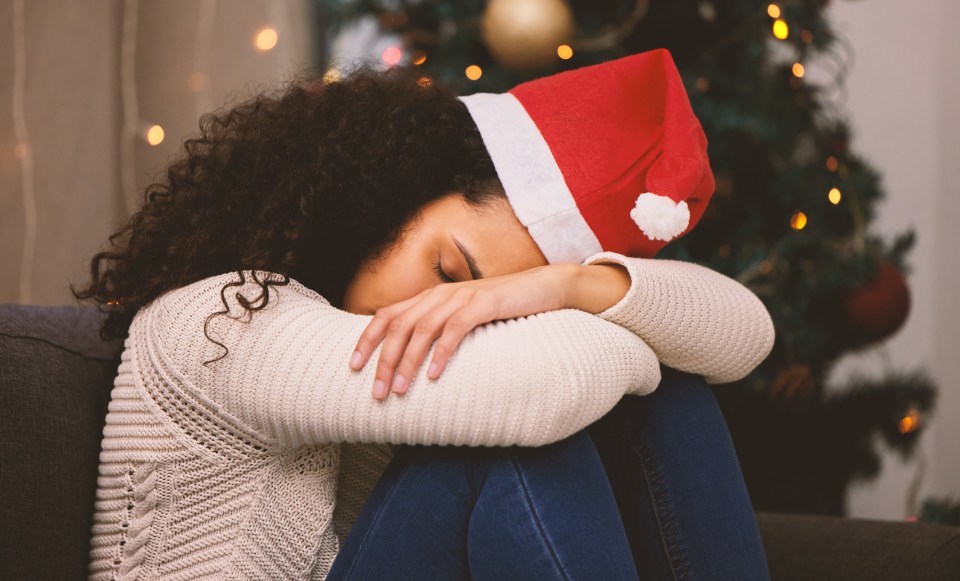 A young woman wearing a Santa hat and a white sweater curls up in a ball on a couch with a blurry Christmas tree in the background.