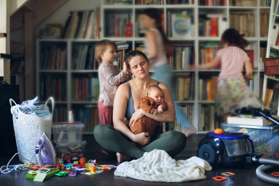 Stressed mother at a table with a laptop, holding an infant while an older child draws next to her.