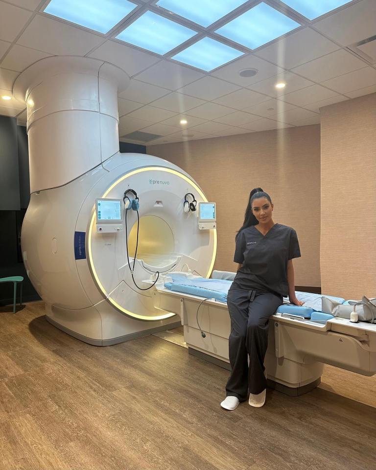 Kim Kardashian wearing gray scrubs, sitting on the bed of a Prenuvo full-body MRI machine.