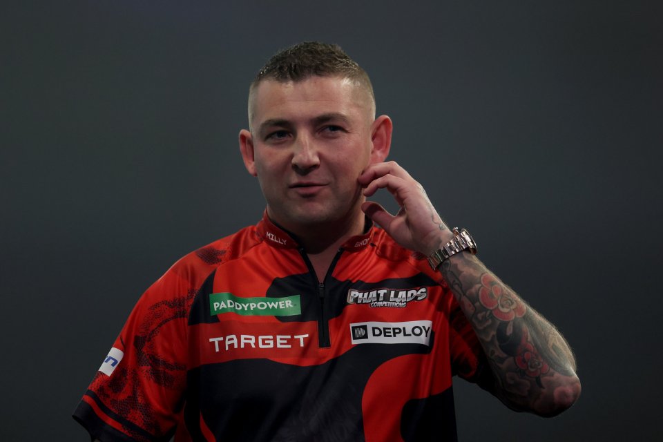 A man in a red and black shirt with sponsors and a tattooed arm touching his ear.