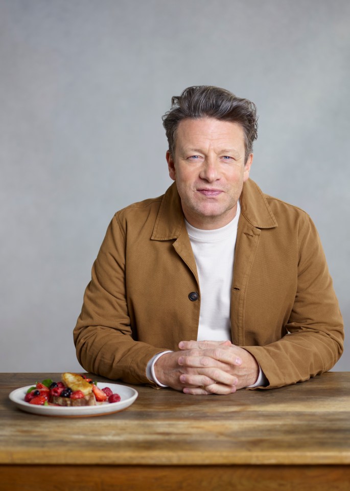 Jamie Oliver sits at a table with a plate of berry French toast in front of him.