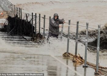 Thousands of California residents found themselves under evacuation orders on Christmas Eve, as disastrous floods and debris flows closed down roads. A man is pictured here hanging on to the railing for dear life after stepping into the path of a rushing culvert on Wednesday