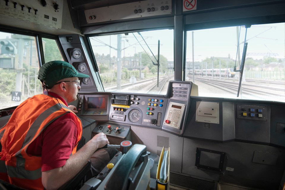 Train engineer in a locomotive, viewed from behind, looking out the front window at the train tracks.