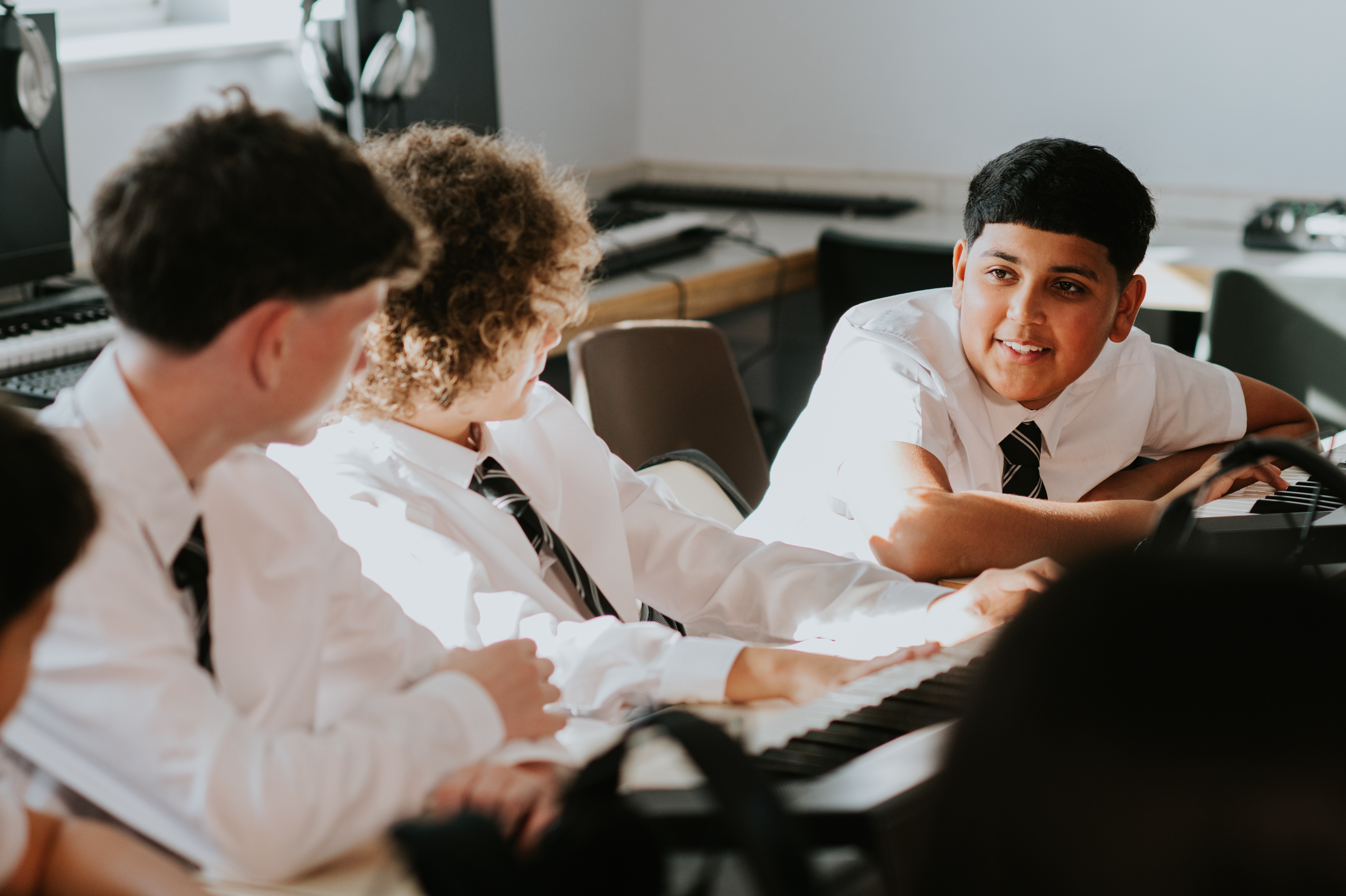 Four high school boys in uniform chat while sitting in a row in a sunny music classroom.