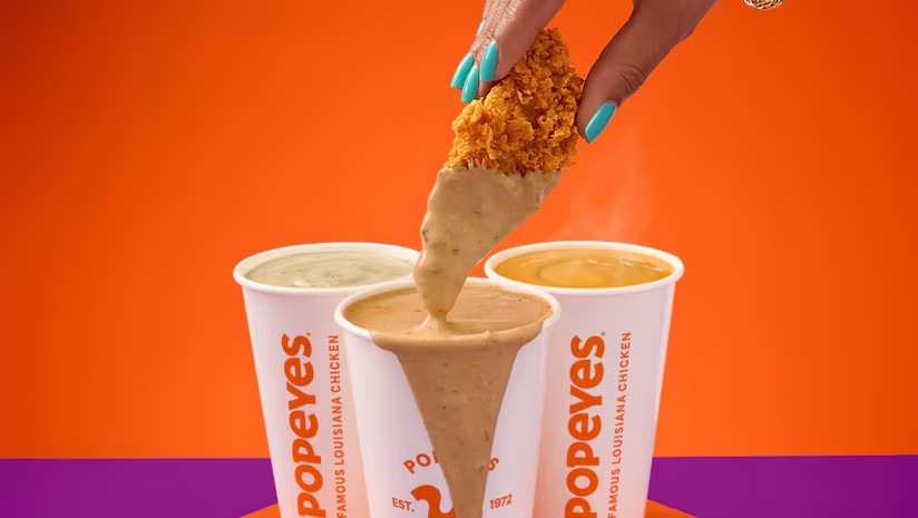 A hand with turquoise nails dips a fried chicken tender into a cup of Popeyes gravy, with two other gravy cups nearby.