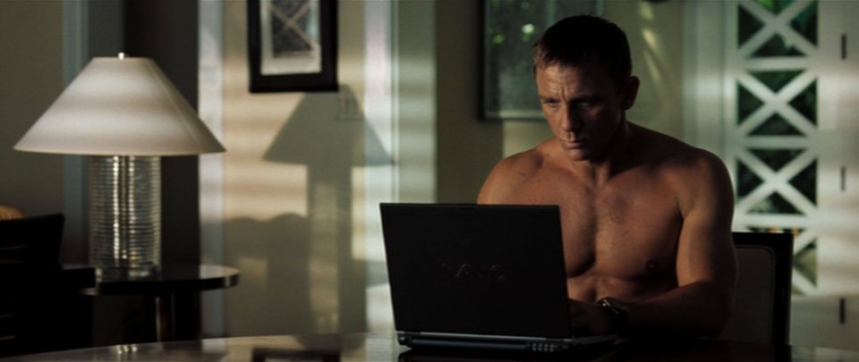 Daniel Craig as James Bond, shirtless, using a laptop.