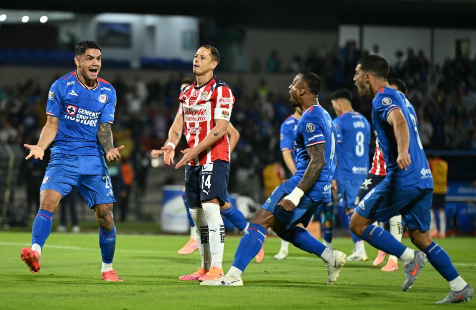 A soccer player in a blue uniform with arm tattoos shouts on the field while another player in a red and white striped uniform looks up with a resigned expression.