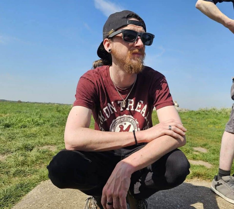 Man wearing sunglasses and a red shirt, crouching outdoors in a grassy field.