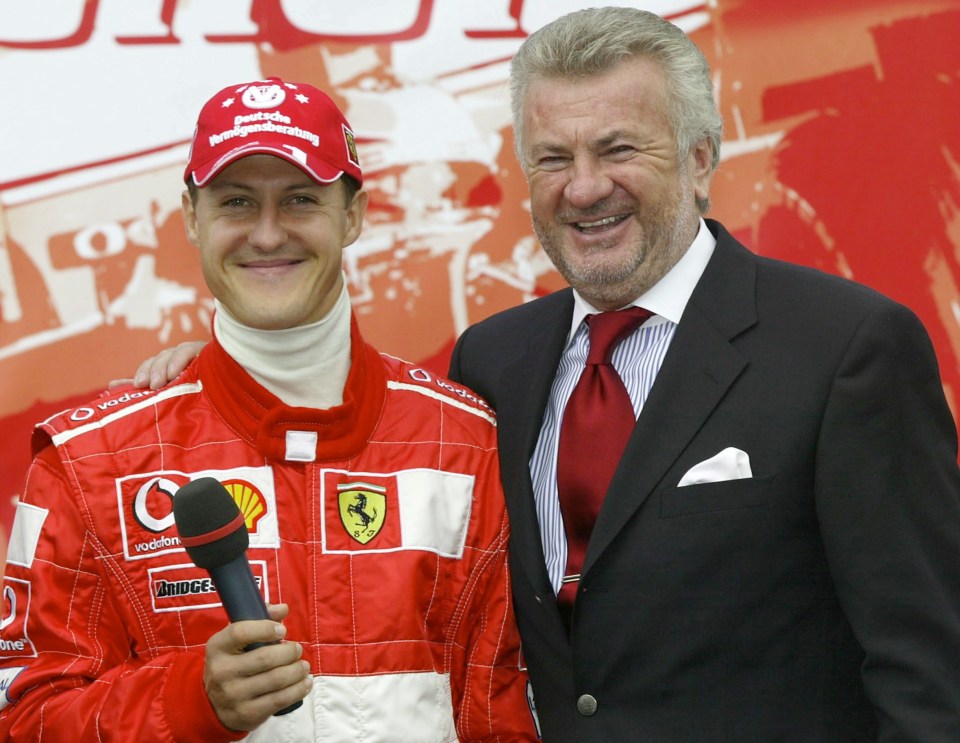 Michael Schumacher in a red racing suit and red cap holding a microphone, with Willi Weber in a suit and red tie.