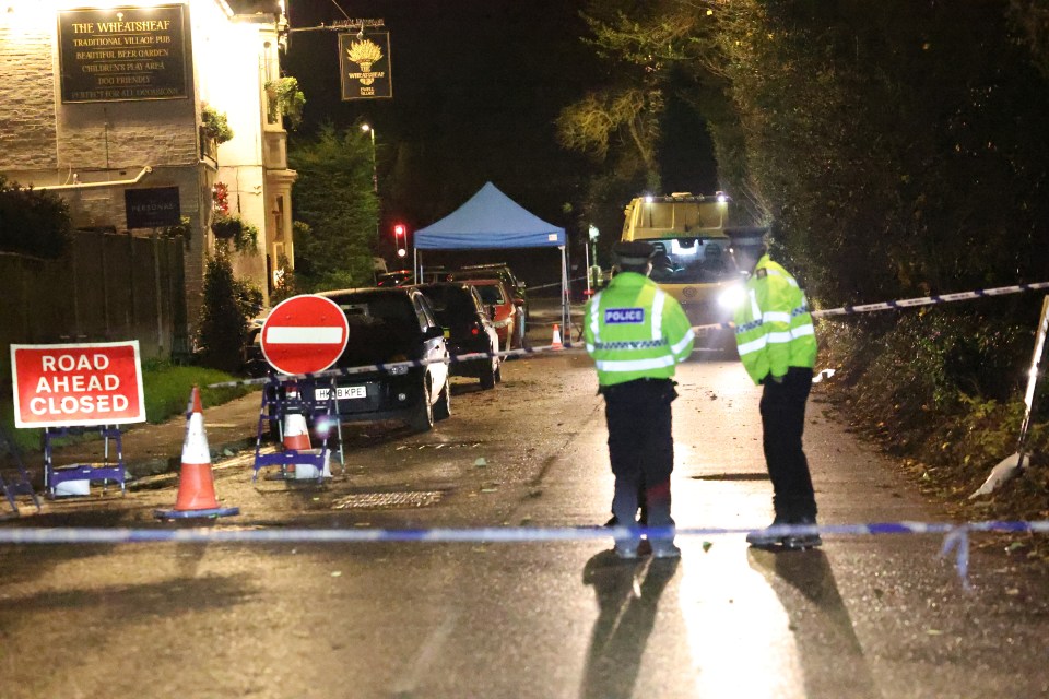 Police tape and a "ROAD AHEAD CLOSED" sign block access to an alleyway near The Wheatsheaf pub, where two police officers stand, investigating a murder.