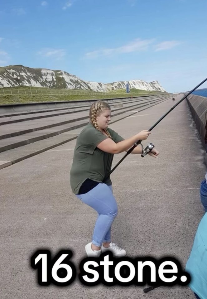 Robyn Quinn, a woman with braided hair, wearing a green shirt and blue jeans, holds a fishing rod while kneeling on a concrete pier with white cliffs in the background.