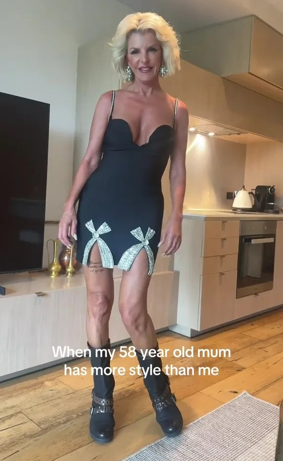 A woman in a black mini dress with sparkly bows and black boots stands in a kitchen.