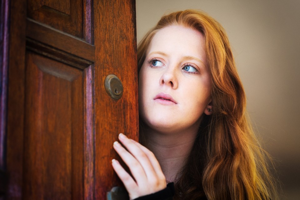 Young woman with red hair cautiously opening her front door.