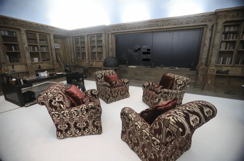 A room with ornate wooden bookshelves, a black desk, a chalkboard with text, and four armchairs with red pillows.