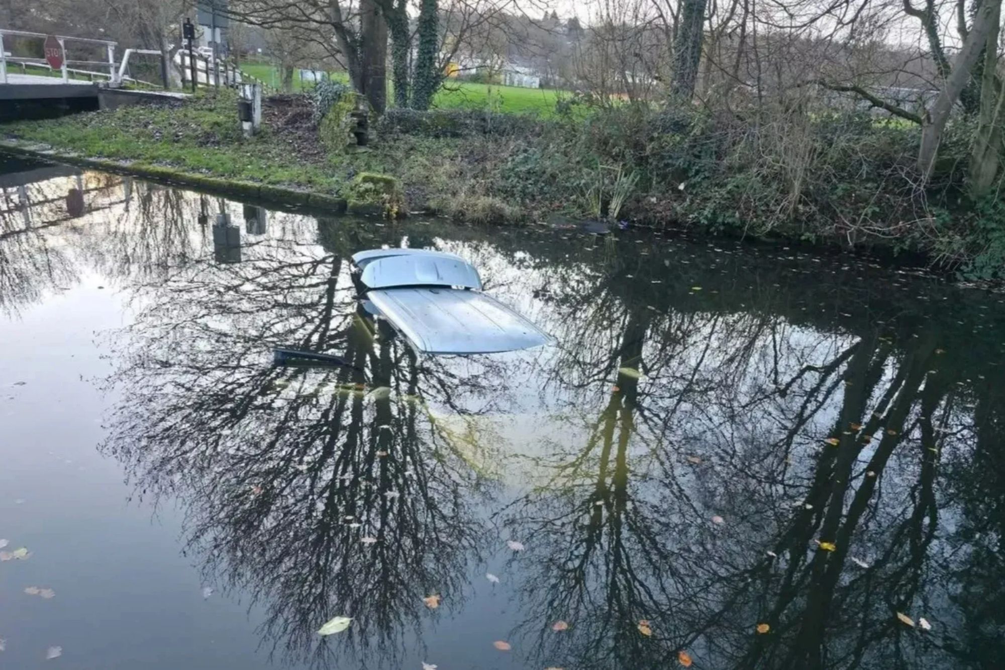 An image collage containing 1 images, Image 1 shows A silver Range Rover Evoque partially submerged in a canal, with tree reflections on the water's surface