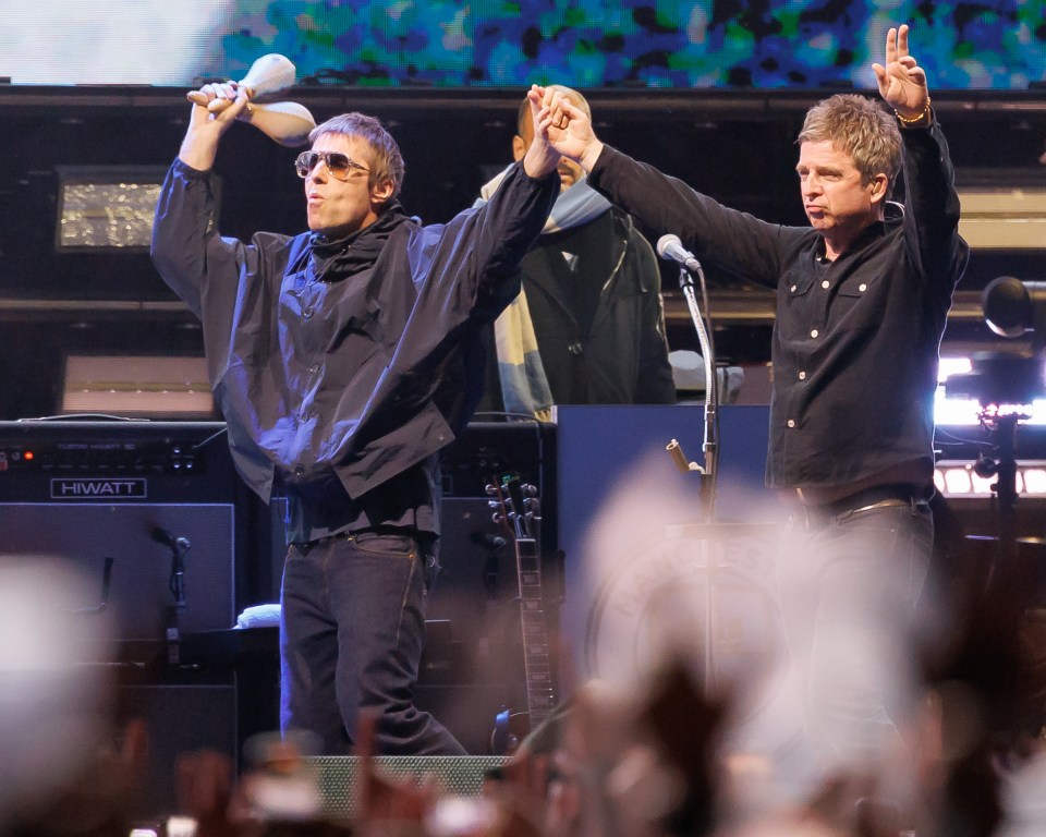 Liam Gallagher holding maracas aloft while Noel Gallagher holds his hands up at an Oasis concert.