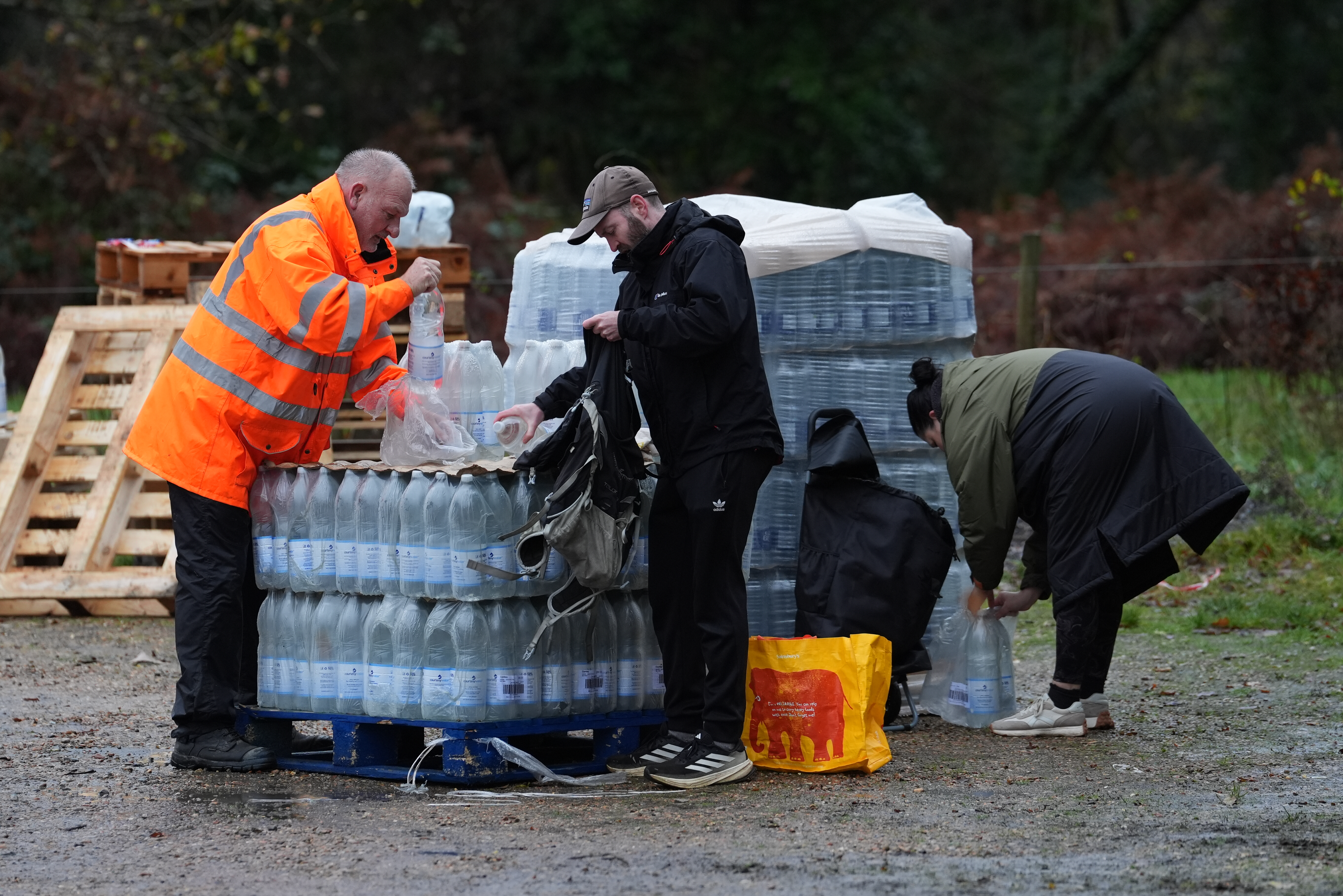 A worker in an orange high-visibility jacket hands out bottled water to people from a stack of palettes.