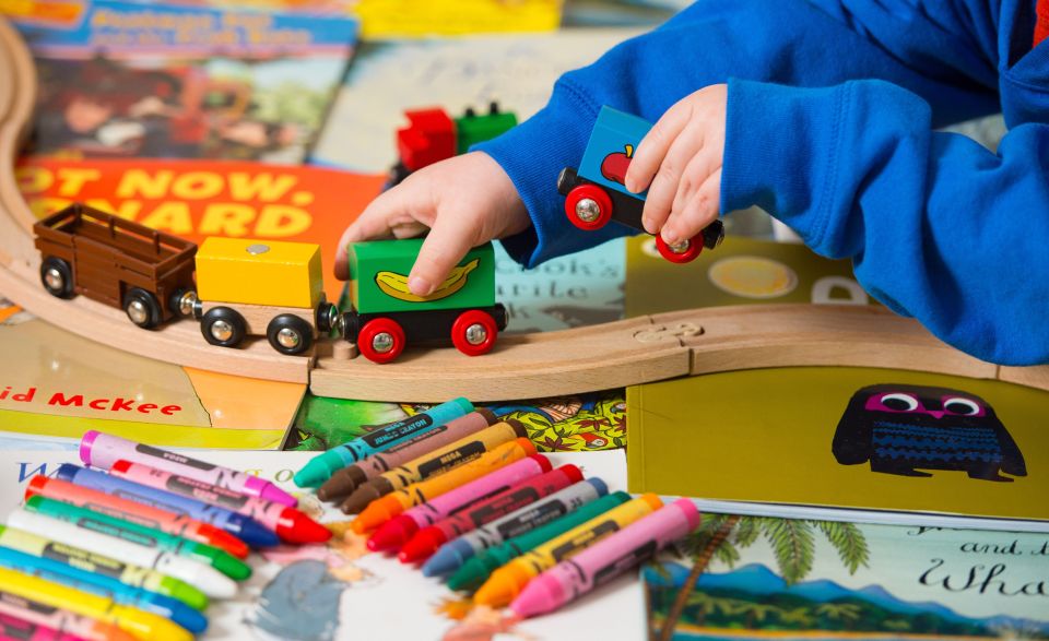 Child's hands playing with a wooden toy train set and crayons on top of children's books.