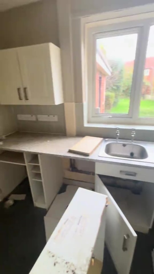 A messy kitchen with an uninstalled cabinet door, an open cupboard, a sink, and a view of a garden through a window.