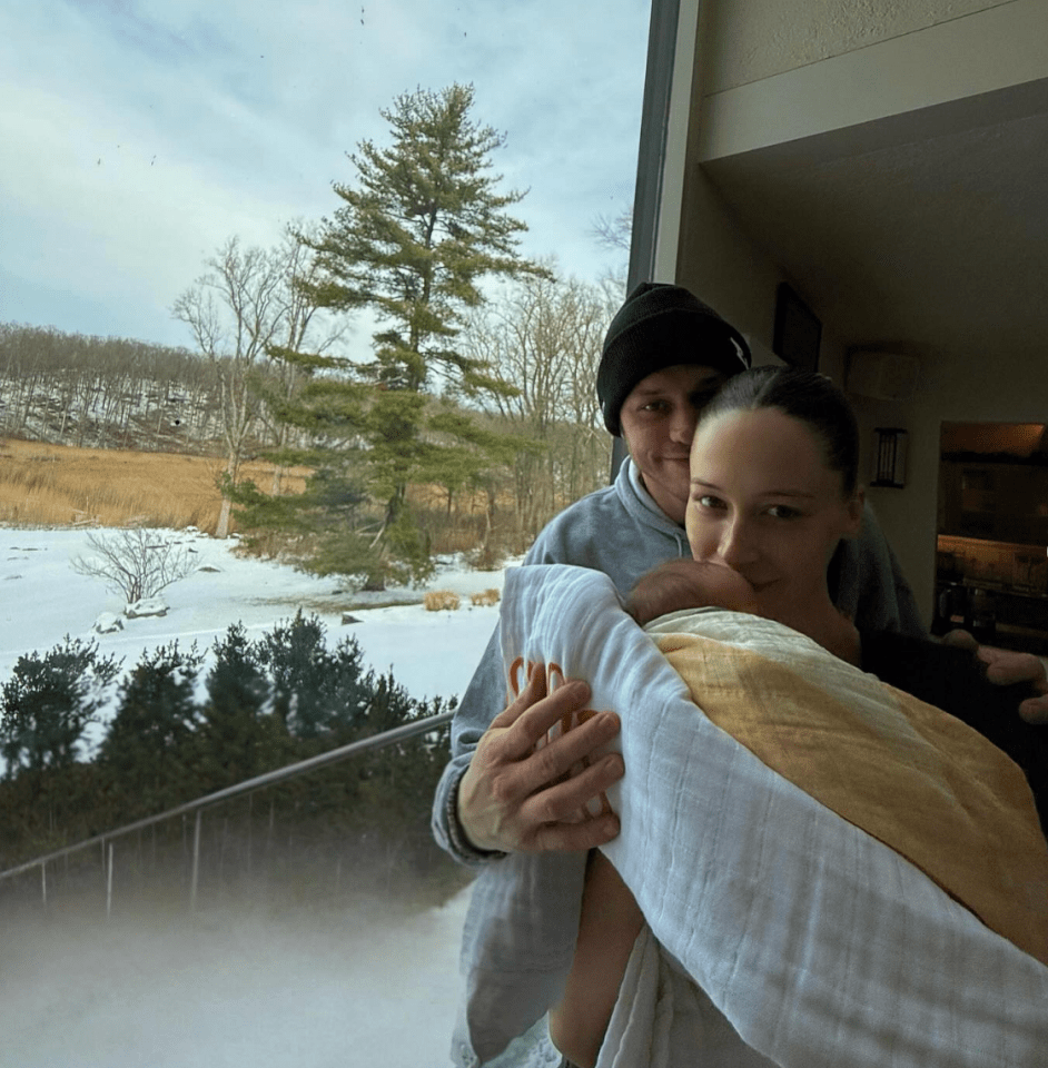 Man and woman holding a baby in a blanket, looking at the camera, with a snowy landscape visible through a window behind them.